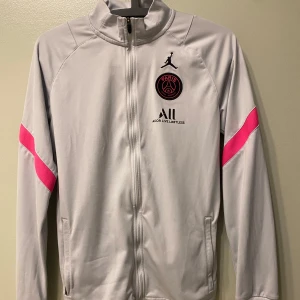 PSG x Jordan grå ziptröja med rosa detaljer - Säljer en vit ziptröja från Jordan x PSG med svarta och rosa loggor på bröstet och rosa detaljer på ärmarna. Tröjan har hel dragkedja, hög krage och är tillverkad i mjukt polyester-material. Perfekt för dig som gillar sportig och clean stil.