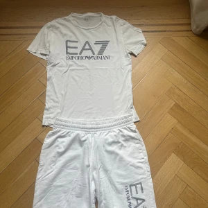 Vit EA7 Emporio Armani T-shirt + shorts  - Vit t-shirt och shorts från EA7 Emporio Armani med stor logga framtill i grått. Tillverkad i mjuk bomull för en skön och avslappnad känsla. Perfekt för en clean och sportig look.