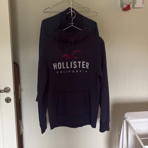  Hollister hoodie  - Säljer en mörkblå hoodie från Hollister med stor broderad logga och text på bröstet. Tröjan har huva med snörning och en klassisk känguruficka framtill. Perfekt för dig som gillar en avslappnad och sportig stil.