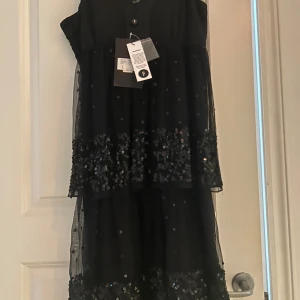 Svart glittrig klänning Sisters Point - Supersnygg svart klänning från Sisters Point med glittriga paljetter på flera lager. Klänningen har ärmlös design och v-ringad hals. Materialet är lätt och har en mesh-känsla med extra volym i kjolen. Perfekt för att sticka ut med coola detaljer.
