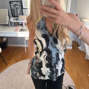 Mönstrad halterneck topp i satin - Säljer en snygg halterneck topp med abstrakt mönster i svart, beige och blå toner. Toppen har knyt i nacken, djup v-ringning och öppen rygg. Materialet är glansigt och känns lyxigt, perfekt för att sticka ut. Passformen är normal och toppen är ärmlös.