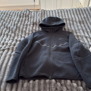 Svart Nike hoodiejacka med dragkedja - Svart hoodiejacka från Nike med hel dragkedja och huva. Jackan har en diskret Nike-logga på bröstet och en snygg vågig detalj över bröstet. Tillverkad i mjukt material, perfekt för chill eller träning. Långärmad och sportig look.