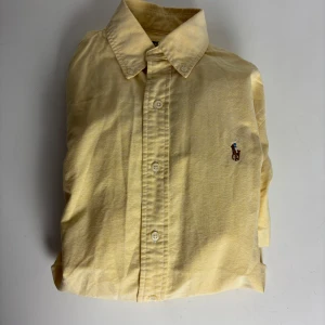 Gul skjorta från Ralph Lauren - Klassisk gul skjorta från Ralph Lauren med broderad logga på bröstet. Skjortan har lång ärm, knappar framtill och en snygg krage. Tillverkad i mjuk bomull som känns skön mot huden. Perfekt för dig som gillar stilrena och tidlösa plagg.