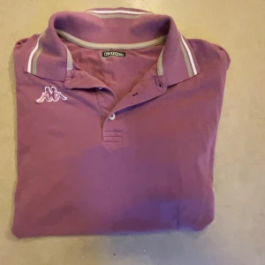 Lila Kappa polo t-shirt med krage - Snygg lila polo t-shirt från Kappa med klassisk krage och vita ränder på kragen och ärmsluten. T-shirten har korta ärmar och Kappa-logga broderad på bröstet. Perfekt för dig som gillar sportig och avslappnad stil.