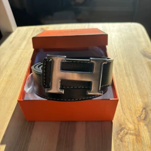 Svart bälte med silver spänne - Säljer ett svart bälte. Bältet är gjort i exklusivt skinn med vit kontrastsöm och levereras i original orange box. Perfekt accessoar för att lyfta vilken outfit som helst.