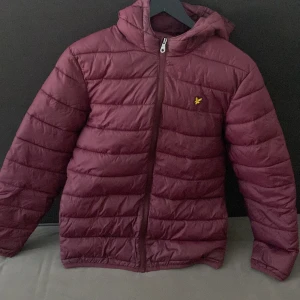 Vinröd pufferjacka från Lyle & Scott - Säljer en vinröd pufferjacka från Lyle & Scott Junior med huva och dragkedja. Jackan har det klassiska gula fågel-loggan på bröstet och är quiltad för extra värme. Perfekt för kyliga dagar och har en snygg, sportig look.