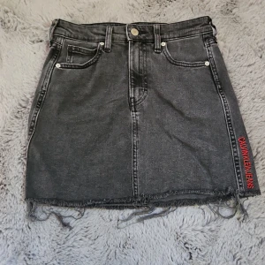 Svart jeanskjol från Calvin Klein - Cool svart jeanskjol från Calvin Klein Jeans med rå kant nertill och röda broderade loggan på sidan. Klassisk femficksmodell, bälteshällor och dragkedja fram. Snygga bakfickor med dekorativ söm. Perfekt till sneakers eller boots. Storlek w25.