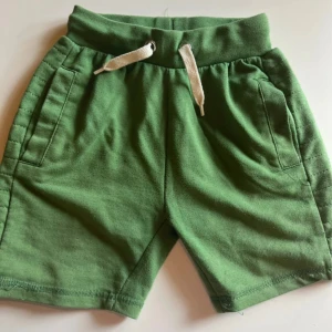 Gröna shorts med snörning, stl 110/116 - Snygga gröna shorts med bred resår i midjan och vit snörning framtill. Mjukt bomullsmaterial och två fickor på sidorna. Perfekta för varma dagar och chill häng.