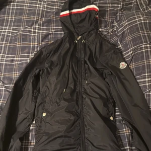 Svart vindjacka från Moncler  - Säljer en svart vindjacka från Moncler storlek 2   med huva och dragkedja. Jackan har Moncler-logga på ärmen och snygga detaljer med röd och blå på luvan  säljes billigt pga ett hål vid armen  men är inget som märks vid användning