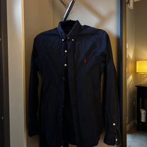Mörkblå skjorta från Ralph Lauren - Klassisk mörkblå skjorta från Ralph Lauren med röd broderad logga på bröstet. Skjortan har knappar framtill och på kragen samt långa ärmar med manschettknappar. Perfekt för en stilren look. Nypris 1599kr