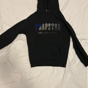 Trapstar svart hoodie med brodyr - Svart hoodie från Trapstar med broderad logga i blått, grått och silver på bröstet. Klassisk känguruficka, snörning i huvan och mjukt material. Perfekt för dig som gillar streetwear och vill sticka ut med en fet logga.