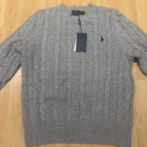 Grå kabelstickad tröja Polo Ralph Lauren - Kabelstickad grå tröja från Polo Ralph Lauren med rund halsringning och klassisk broderad logga på bröstet. Tröjan har ribbade muddar och är perfekt för lager-på-lager under kyliga dagar. Snygg och tidlös design som funkar till allt.