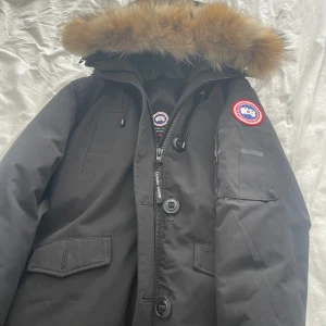 Svart parkajacka från Canada Goose - Dam jacka Priset går o diskutera. Säljer en svart parkajacka från Canada Goose med ikonisk patch på ärmen och stor huva med äkta päls. Jackan har dragkedja, knappar och flera praktiska fickor. Perfekt för kalla vinterdagar och riktigt snygg streetstil.