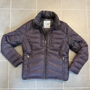 Mörkblå dunjacka från Zegna - Snygg mörkblå dunjacka från Zegna med quiltad design och hög krage. Jackan har två dragkedjeförsedda fickor framtill och en stilren look. Perfekt för kalla dagar med lätt och värmande känsla. Klassisk passform och diskret logga på insidan. Storlek M.