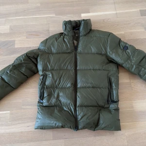 SNOOT JACKA vinter - Säljer en mörkgrön pufferjacka från Sail Racing med hög krage och två sidofickor med dragkedja. Jackan har ett stilrent, quiltat mönster och långärmade armar. Perfekt för kalla dagar och har en snygg, modern look. Luva tillkommer också