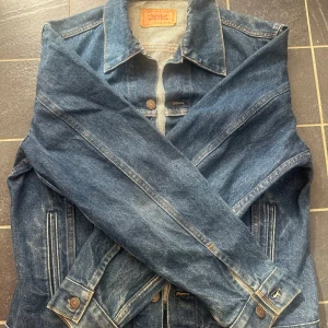 Levi's klassisk jeansjacka blå - Säljer en klassisk blå jeansjacka från Levi's med snygga bronsfärgade knappar och två bröstfickor med lock. Jackan har normal passform, krage och långa ärmar. Perfekt för dig som gillar tidlös streetstil och vill ha en jacka som funkar året runt.