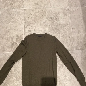 Olivgrön stickad tröja J.Lindeberg - Snygg olivgrön stickad tröja från J.Lindeberg med rund halsringning och diskret broderad logga på bröstet. Långärmad modell i mjukt material som passar perfekt till lager-på-lager. Enkel och stilren design som funkar till allt. Storlek S, pris kan ändras vid köp!