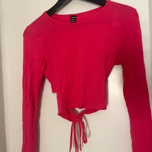 Rosa croppad topp med knyt från SHEIN - Trendig rosa långärmad croppad topp från SHEIN med rund halsringning och snygg knytning baktill. Tunn och lätt i materialet, perfekt för att skapa en cool och avslappnad look. Passar dig som gillar att sticka ut med färg och detaljer.