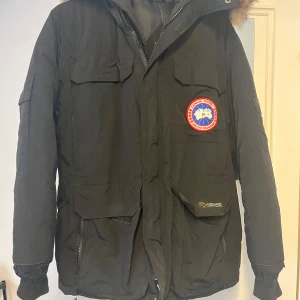 Svart dunjacka från Canada Goose - Canada Goose dunjacka i svart med ikonisk röd och blå Arctic Program-patch på bröstet. Jackan har flera stora fickor, huva med pälsdetalj och dragkedja framtill. Tillverkad i nylon med fyllning av dun och fjäder, perfekt för kalla vinterdagar. Står storlek XS fast jag skulle säga att den passar M-L. De ändå defekterna är att innerfickan är lite trasig och delen av dragkedjan som man drar i har knäckts i hälften. Mått: 📏   Armhåla till armhåla: ≈ 60cm (rakt över) Midja: ≈ 50cm (rakt över) Jackans