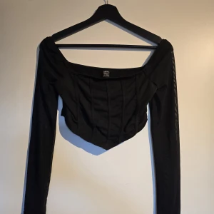 Svart långärmad korsett-topp - Svart korsett-topp med långa meshärmar och markerade sömmar framtill. Toppen har offshoulder-skärning och croppad passform som ger en edgy vibe. Perfekt för dig som vill sticka ut med en stilren och trendig look. Endast testad