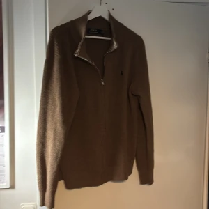 Brun stickad kofta från Polo Ralph Lauren - Säljer en brun stickad kofta från Polo Ralph Lauren med blå broderad logga på bröstet. Koftan har hel dragkedja, hög krage och långa ärmar. Perfekt för lager-på-lager och chill höst- eller vinterstil.