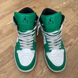 Nike Air Jordan 1 Mid Grön/Vit - Säljer ett par Nike Air Jordan 1 Mid i grön och vit färg med klassisk svart logga på sidan och tungan. Skorna har snörning, svart innersula och vit/grön ovandel i skinn. Perfekt för dig som vill sticka ut med en ikonisk sneaker.