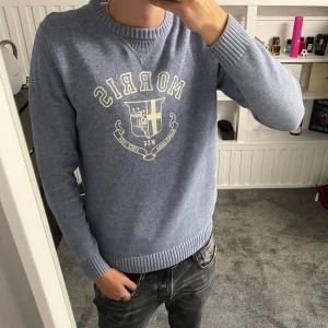 Blå stickad tröja från Morris - Snygg blå stickad tröja från Morris med tryckt logga och emblem på bröstet. Tröjan har rund halsringning, ribbade muddar och är långärmad. Perfekt för dig som gillar klassisk och stilren. 