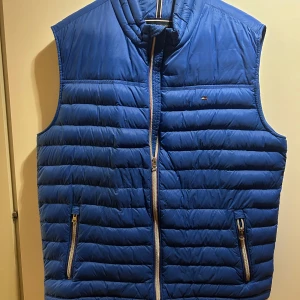 Blå dunväst från Tommy Hilfiger - Snygg blå dunväst från Tommy Hilfiger med quiltad design och två dragkedjefickor framtill. Västen har ståkrage och klassisk logga på bröstet. Perfekt för lager-på-lager och enkel att matcha med hoodies eller tröjor.