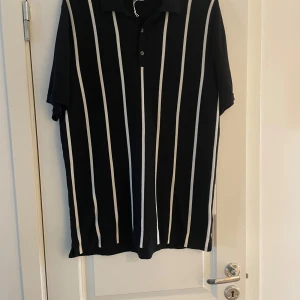 Svart randig pikétröja från H&M - Svart pikétröja från H&M i premium bomull med vita vertikala ränder. Klassisk krage, knäppning med två knappar och korta ärmar. Snygg och stilren modell som passar perfekt till jeans eller shorts.