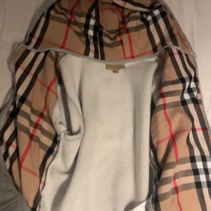 Grå Burberry hoodie med klassiskt foder - Grå hoodie från Burberry med dragkedja och snörning. Storlek S. Perfekt skick. Insidan av huvan har det ikoniska rutiga Burberry-mönstret i beige, svart, rött och vitt. Tröjan har fickor framtill och diskret Burberry-logga på bröstet. Perfekt för dig som vill ha en stilren och lyxig look.