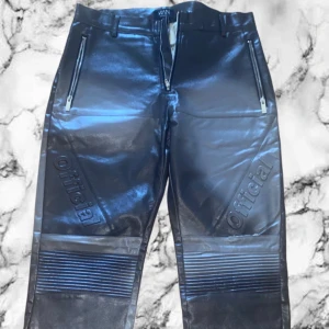 Svarta skinnbyxor med dragkedjor - Snygga svarta skinnbyxor från BOHOOMAN Official med coola detaljer som dragkedjor vid fickorna och präglad text 'Official' på benen. Byxorna har ribbade partier över knäna för en extra edgy look. Perfekta för dig som vill sticka ut med en streetstyle vibe.