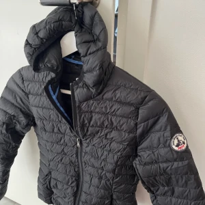 Svart dunjacka med huva från JOTT - Svart quiltad dunjacka från JOTT med huva och dragkedja framtill. Jackan har en smidig passform,  logga på ärmen och blå detaljer på insidan. Perfekt för kyliga dagar när du vill hålla stilen enkel och clean.