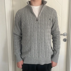 Grå stickad half zip från Polo Ralph Lauren - Säljer en grå kabelstickad tröja från Polo Ralph Lauren i storlek XL. Tröjan har half zip. Är xl i barn storlek men som s i vuxen.