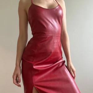 Röd lång klänning i skinnimitation - Säljer en röd lång klänning i skinnimitation med smala axelband som korsas i ryggen. Klänningen har en tight passform och en snygg slits framtill som ger en cool och edgy look. Perfekt för dig som vill sticka ut med en statement piece.