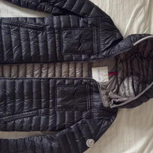 Svart Moncler dunjacka med huva ( moncler Clovis 2017)  - Snygg svart dunjacka från Moncler med grått foder och huva. Jackan har två bröstfickor med lock, dragkedja framtill och klassisk Moncler-logga på ärmen. Lättviktig och quiltad design som ger en clean och modern look. Priset kan diskuteras. 