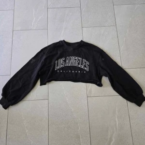 Svart croppad sweatshirt Los Angeles - Svart croppad sweatshirt med trycket 'Los Angeles California' framtill. Tröjan har långa ärmar med muddar och en avslappnad passform. Perfekt för en chill och trendig look.