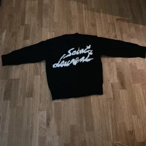 Svart Saint Laurent stickad tröja - Svart stickad tröja från Saint Laurent med vit, fluffig logga framtill. Tröjan har rund halsringning och långa ärmar. Perfekt för dig som vill ha en statement piece med lyxig känsla och cool design.