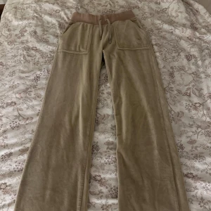 Beiga mjukisbyxor Juicy Couture XXS - Supermysiga beiga mjukisbyxor från Juicy Couture i velour med bred resår och snörning i midjan. Byxorna har raka ben, stora fickor fram och bak med knappdetaljer. Perfekta för chill dagar och har den klassiska Juicy-looken. De har små defekter vilket du kan se på de sista bilderna!