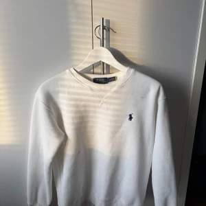 Vit sweatshirt från Polo Ralph Lauren - Vit sweatshirt från Polo Ralph Lauren med klassisk rund hals och broderad marinblå logga på bröstet. Tröjan har långa ärmar och ribbade muddar vid ärmslut och nederkant. Perfekt för en clean och stilren look.