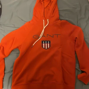 Orange hoodie från GANT med tryck - Snygg orange hoodie från GANT  med vit snörning i huvan och stort tryck med logga och sköld på bröstet. Klassisk modell med lång ärm och mjukt material, perfekt för chill dagar. Passar dig som gillar streetwear och vill sticka ut lite extra. Priset är inte hugget så det är bara att komma med bud! Den är helt ny!