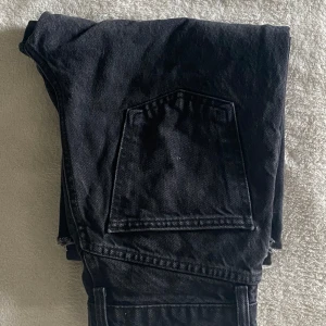Svarta wide jeans från Zara - Svarta jeans från Zara med bred passform och raka ben. Jeansen har stora bakfickor, råa kanter vid benslut och klassiska detaljer som nitar och knappstängning. Tillverkade i kraftigt denimtyg som ger en cool och avslappnad look.