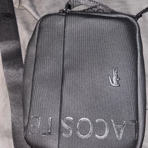 Svart axelväska från Lacoste - Snygg svart äkta axelväska från Lacoste med justerbart band där Lacoste-loggan är invävd. Väskan har en stilren design med präglad krokodil på framsidan och dragkedja både fram och bak. Tillverkad i slitstark syntet med en clean look. Även oanvänd