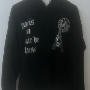 Rhinestone Hoodie - Svart hoodie med huva och coolt Paris-tema i strass på framsidan. Texten 'Paris au clair de lune' och motiv av Eiffeltornet och måne i glittriga stenar ger en unik vibe. Perfekt för dig som gillar statement-plagg och snygga detaljer.