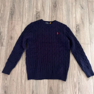 Mörkblå stickad tröja Polo Ralph Lauren - Mörkblå kabelstickad tröja från Polo Ralph Lauren med klassisk röd logobrodyr på bröstet. Rund hals och ribbade muddar vid ärmslut och nederkant. Perfekt för dig som gillar stilren och tidlös design.