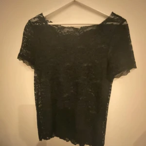 Svart spetsblus med kort ärm - Säljer en svart blus i genombruten spets med korta ärmar. Toppen har en rak passform och vacker blommig spets över hela plagget. Perfekt för dig som vill ha en stilren men ändå detaljrik topp. Snygg att styla med både jeans och kjol.