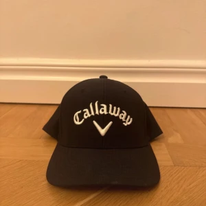 Svart keps Callaway Odyssey - Svart keps från Callaway med broderad vit logga framtill och Odyssey-detalj bak med röd cirkel. Justerbar rem baktill med logotyp. Klassisk böjd skärm och ventilerande mesh på insidan. Perfekt för golfbanan eller vardagslooken.