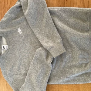 Grå crewneck från Nike XS - Snygg grå crewneck från Nike i klassisk modell med rund hals och lång ärm. Tröjan har en broderad vit Nike-logga på bröstet och är gjord i mjuk bomullsblandning. Perfekt för en chill och sportig look.
