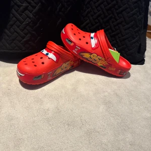 Röda Crocs Lightning McQueen 42-43 - Säljer ett par röda Crocs Crocband med Lightning McQueen-motiv från Disney Cars. Skorna har coola detaljer som ögon och nummer 95 på sidorna. Tillverkade i syntetmaterial med ventilerande hål och justerbar hälrem. Perfekt för dig som vill sticka ut med en lekfull stil. Helt nya. Nypris 799kr