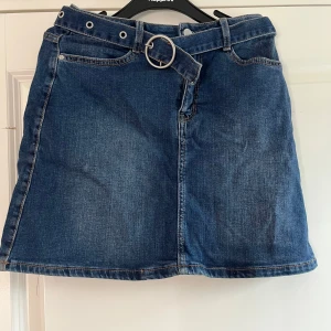 Jeanskjol med bälte  - Mörkblå jeanskjol  med snyggt bälte och silverfärgat spänne. Kjolen har klassiska jeanssömmar, fickor fram och en rak, kort passform. Perfekt för dig som gillar en enkel men trendig look.