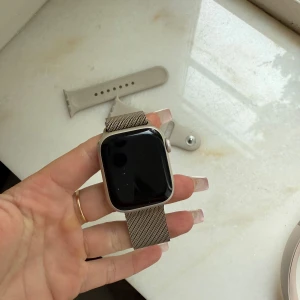 Apple Watch med silver mesh-armband - Snygg Apple Watch med rektangulär svart skärm och ett stilrent rosé mesh-armband i metall. Klockan har en modern design och det medföljer även ett extra ljusbeige silikonarmband. Perfekt för dig som gillar tech och vill ha koll på tiden med stil.